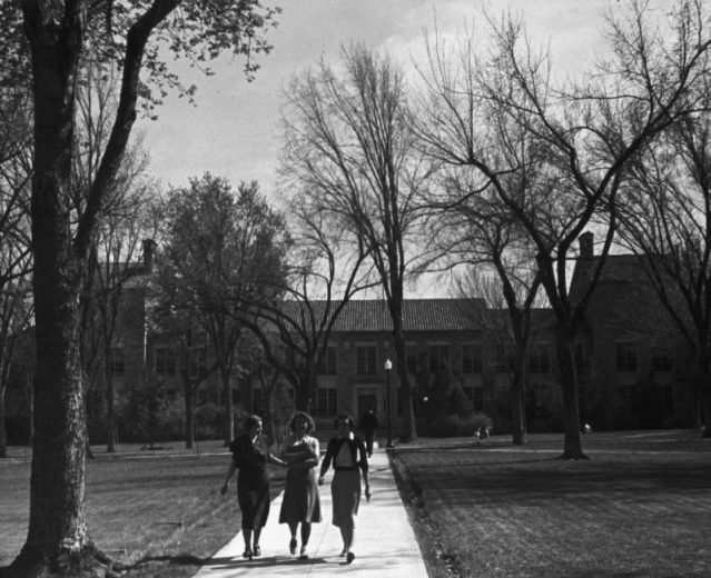 Gabled, three-story wings flank the central section of the arts building at the University of Colorado at Boulder. In architect Charles Z. Klauder's rural Italian motif, the sandstone edifice has a gabled, terra- cotta roof with chimneys. Stone entablature finishes the windows and doors. Trees, lawn, sidewalks and a cast iron street lamp landscape the grounds, and three women approach, wearing sweaters and carrying books.