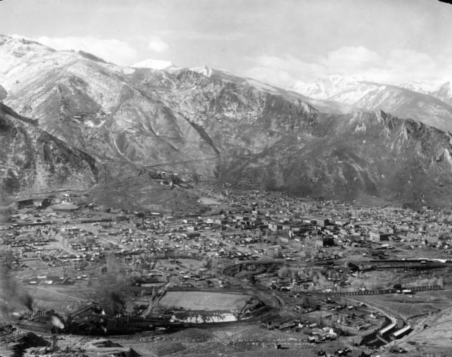 Aerial view of Aspen (Pitkin County), Colorado. Denver and Rio Grande Railroad tracks wind past industrial smoke stacks near the Smuggler, Silver King, and Mollie Gibson Mines. Prominent buildings in town include St. Mary's Church, and the Wheeler Opera House. The Sampler Mine, and Argentum-Juniata mines are on the slopes of West Aspen (Shadow) Mountain in the distance.
