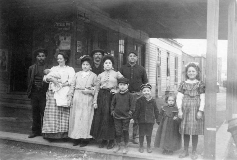 A group of Italian families pose near a shop in a coal camp probably in Las Animas County, Colorado. One woman holds a baby in her arms and smiles. The children, dressed in sweaters, coats, and dresses also smile.  The men stand behind the women.