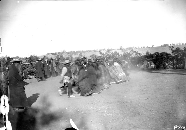 Native American men and women take part in a Bear Dance near Ignacio, La Plata County, Colorado.  A line of men face a line of women while groups of white and Native American men, some standing near a wooden pole and brush fence, all wearing hats, look on.