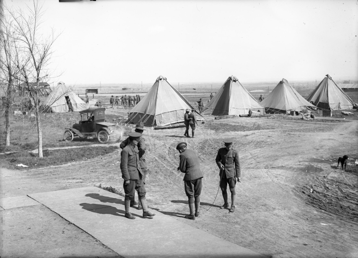View of camp life at Fort Logan, Colorado, a World War I assembly  point for young soldiers, includes four sided pyramidial canvas tents, a automobile and a dog. Officers wearing uniform gather in foreground.
