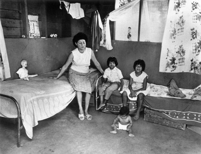 A Hispanic woman sits on a bed with her children at her house in Colorado. A doll and jacket are on the beds. Curtains and clothes on hangers hang from the walls nearby.