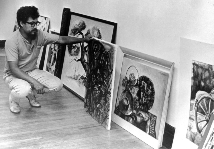 Mexican American artist Carlos Santistevan kneels down to look at a painting possibly in the Centro de la Cruzada in Denver, Colorado. Additional paintings are on the floor and lean against the wall. Carlos Santistevan was the founder of the El Grito Gallery in Denver.
