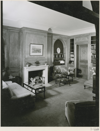 Interior view of a den in the Whiteman House in the Harvey Park neighborhood of Denver, Colorado. A two-story addition to this house was designed by architect Burnham Hoyt in the 1920s for Henry de Compiegne. The sitting room has wood paneled walls with moldings and a chair rail. Walls have built-in bookshelves and arched niches. The carpeted room has a couch, armchairs and ottomans. Shows a glass topped coffee table, flowers on side tables, and a horse and soldier sculpture on the fireplace mantel.