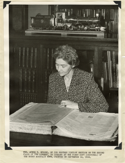 Agnes Wright Spring, sits at a table in the Western History Department of the Denver Public Library and looks at the first copy of the Rocky Mountain News. Behind her are wood bookcases and the 1:16 scale model of Colorado Midland No.9 engine and tender made by Claude E. Harris (1938).