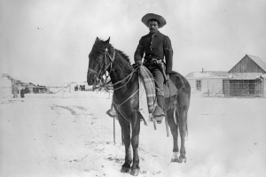 X-31380, A Black Corporal, (Buffalo Soldier) in the Ninth Cavalry, poses on his horse on the Native American (Lakota Sioux) Pine Ridge Agency, South Dakota. He wears a military uniform and has a gun in a holster on his belt. Snows covers the ground and frame agency buildings show in background.