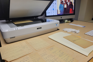 An epson scanner connected to a mac desktop, archival papers are laid out on the desk, a pair of white gloves are used to handle the archival papers.