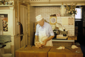 Portrait of Audi Cerrone, butcher and proprietor of Cerrone's Italian Market at 3617 Osage Street in the Highland Neighborhood of Denver. He stands behind a butcher block and holds a cut of meat. Shows the door to a walk in freezer, and a statue of the Virgin Mary, and a vase of dried flowers. Poster and certificates are on a wall.