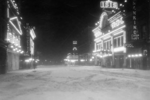 Night view of Curtis Street in Denver, Colorado; shows snow and movie theaters. Electric signs read: "Marquette," "Iris Theater," "Isis," and "The Nanking Chop Suey, Pool."