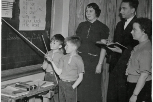 Photograph of two unidentified students, and three staff members from University Park Elementary School located in Denver, Colorado.  In the picture, two boys stand to the side of the blackboard.  They have pointers in their hands.  Two women and one man stands behind the blackboard.  The man appears to be writing something in a notebook.