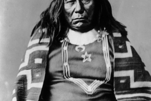 Portrait of Chief Colorow, called Colorado, Native American Ute Indian; he wears long hair with banks tied back in a fur tail and is wrapped in a woven patterned blanket.