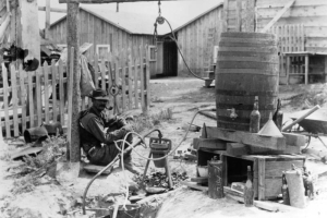 A man smokes a pipe as he sits next to a contraption of some kind, possibly a smelter or liquor still, in (probably) the Bohannon Placer District in Lemhi County, Idaho. Hoses come from a wooden barrel and center on what is perhaps a forge or crucible. Bottles, buckets, scrap lumber and a wheelbarrow lay around the device, with board and batten buildings in the background.