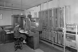 An operator works at a switchboard at the Mountain States telephone exchange building in Limon (Lincoln County), Colorado. Shows telephone equipment and telephone booths on the far end of the room. The woman wears a headset and polka-dot dress.