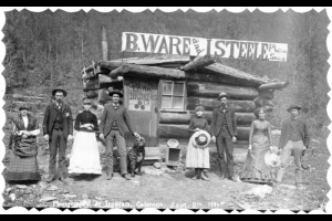 In Ironton, Ouray County, Colorado, people and a dog pose by a log business with signs: "B. Ware and I. Steele. Plain Sewing;" and "Pawn Brokers Co." The men are in suits with gold chains; the women wear flounced  skirts with draped overdresses, knit shawls, and tight bodices.