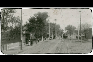Photograph of a street in the Globeville neighborhood of Denver, Colorado.  Pictured is an electric street car in the street, with various individuals walking along the sidewalks. Date estimated based on start of electric trolley service in this area (1908) and street scene.