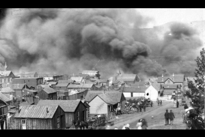 View of April 29th, 1896, fire and clouds of smoke from junction of Myers Avenue and "A" Street after explosion at El Paso Livery stable, Cripple Creek, Colorado; scene includes horse-drawn wagons with household goods or personal belongings parked in front of small wooden frame residences (probably cribs of infamous red-light district of Myers Avenue); men and women are in front of residences and on the dirt street.