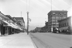View of Broadway at Ellsworth Street, looking north in Denver, Colorado. Multi-story  commercial buildings, street lamps and street railway tracks line the street. Notable buildings include the Broadway Bank building and the Queen Theater. A person rides a bike and people stand near storefront windows and under awnings.  Automobiles are parked nearby. Signs on buildings read: "Restaurant," "William Small, Groceries & Meats," "Coal," "Sorensen Fuel and Feed" and "Queen."
