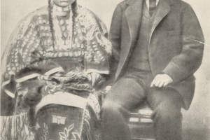Portrait of George Bent, a mixed-blood who chose to live and fight as a Cheyenne warrior, with his wife Magpie. Bent wears a suit and moccasins, he has a moustache. Magpie wears a dress decorated with elk teeth, quillwork and fringe, and a hairpipe choker; her hair is in braids.