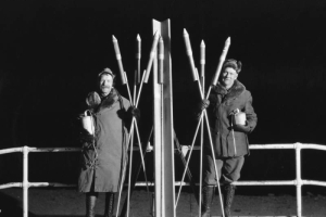 Members Garrett and MacDonald of the AdAmAn Club pose with fireworks including rockets and bombs with fuses, on top of Pikes Peak, Colorado Springs, El Paso County, Colorado. They wear tall lace-up boots and fur collared parkas.