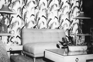 Black and white print of living room from unidentified Utility/Economy Model home in Aurora's Hoffman Heights Development. Small, light colored couch without arms in front of light colored coffee table with plant. End table on either side of couch. Lamps with people as base. Patterned drapes with feathers on it behind couch.