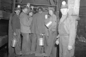 Miners stand in and near the cage of a coal mine in Colorado. They hold lunch pails and wear miner's lights.