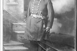 Studio portrait of Major Rafael Chacón (retired). He is dressed in his United States Army uniform. His jackets has two rows of brass buttons, he wears a cap and has a sword at his waist. He stands in front of a painted backdrop.
