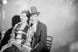 A man and a woman dressed in costume for Saint Patrick's Day pose, probably Denver, Colorado. She wears a dress with ruffled sleeves, lace and satin bodice, and a tiara. He wears a hat and suit with satin and wide lapels.