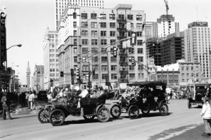 View of the Saint Patrick's Day Parade at 16th (Sixteenth) Street and Broadway in the Central Business District, Denver, Colorado. Men and a woman in hats ride in decorated antique convertible cars. The Paramount Theater and Daniels and Fisher Tower are in the distance.
