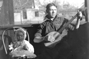 Portrait of a young man playing a banjo in Denver, Colorado, by a baby in a fur hood and lace dress.