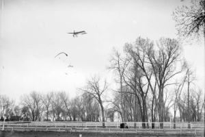 View of a Bleriot airplane in flight over Denver, Colorado, shows trees and fences.