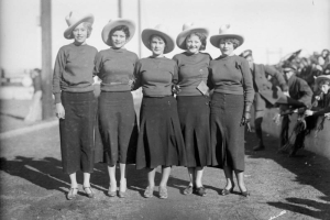 Five University of Wyoming cheerleaders wear matching cowboy hats and sweaters with wool skirts at a football game. One woman wears a tag that reads: "Stop North On Their Victory March.".