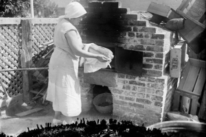 A woman holds a loaf of bread in a towel, by an outdoor brick oven.