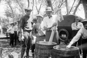 A group of men stand by wooden barrels at a Gates Rubber Company picnic or outing in Colorado, One man uses a wooden paddle while another man uses a cup or a ladle.