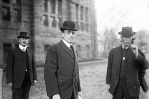 Three men stand outside of the Denver City Jail during the Brown Palace murder trial in Denver, Colorado. They wear suits, vests and hats. One man holds a cigar. The man in the center, with a rolled piece of paper in his hand, is the defendant, Frank Henwood.
