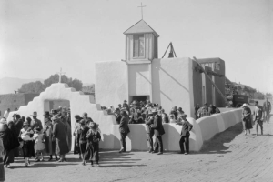 A crowd of men, women, and children exit an adobe church in Taos Pueblo (Native American), New Mexico. The church has a walled courtyard, stepped arch gate with a cross, wood steeple with a cross, and a bell on the roof. A car is parked in back.