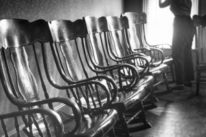 An interior view of the Clear Creek County Courthouse (?) in Georgetown, Colorado, including five Victorian rocking chairs lined up, a woman in slacks and a peasant blouse looking out the window, another rocking chair (partially visible), and wallpaper with small print. The chairs and floor shine from the window's light.