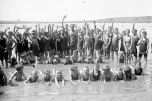 A man in shorts and a tank top with letters: "Chief Life Saver" poses in a crowd of boys and girls wearing swimming suits by a lake.