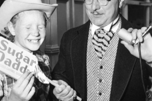 The Mayor of Central City, Colorado, wearing a suit, top hat, and glasses, holds a microphone in his left hand and, with his right, hands a large Sugar Daddy candy bar to a boy wearing a plaid shirt, blue jeans, and a cowboy hat. The boy, whose eyes are sqeezed shut, is smiling. A Central City, Colorado, storefront is in the background.