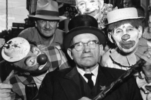 In Central City, Colorado, a man in a suit, top hat and glasses, wearing a pin styled after a sheriff's badge and holding a cane, is surrounded by the faces of three clowns and another man in a hat. The clown  on the right is holding onto the man's cane. The clown above the man has a  ruffled collar and a painted face. The third clown wears a plaid suit, a red rubber nose, and a tiny straw hat.