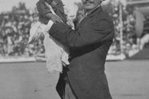 A man poses with a Native American (tribe unknown) infant in the stadium at the Festival of Mountain and Plain. The baby is wearing an oversized shirt and tattered pants. The man is wearing a suit and hat.