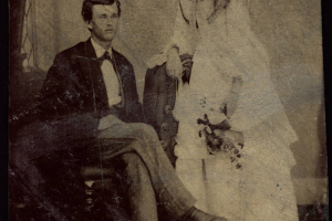 Studio portrait (sitting and standing) of a husband and wife probably on their wedding day. She holds a bouquet and wears a dress with a bustle.