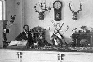 Interior view of the Manitou House Hotel office, in Manitou Springs (El Paso County), Colorado; decor includes taxidermy mounted squirrels, a lynx, a beaver, birds, a rabbit, and deer. A Regulator clock, rifles, pistols, a safe, and a kerosene lamp are on the wall; a man with mutton-chop sideburns rests his arm on the guest book. Men in bowler hats play cards, and a woven runner reads: "Land Office."