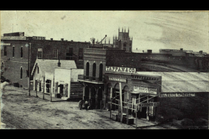 View of 15th (Fifteenth) (F) Street in Denver, Colorado. Signs on the wood and brick business district buildings read: "Broadway Store Dry Goods & Clothing Wholesale & Retail", "Tappan & Co.", "Chemicals Paints Oils Brushes & Glass", and "Drugs". Lawrence Street Methodist Episcopal Church is in the distance.