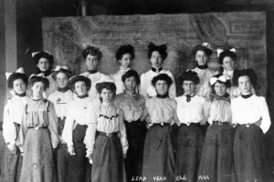 Sixteen members of the Leap Year Club in Basalt, Colorado pose in  two lines. Ten women are in the first row, and six women are in the second row. Characteristic of the early twentieth century, their clothing includes long-sleeved, high-collar blouses with puffed sleeves and long skirts. Several of the women wear jewelry, including earrings, cameo broaches, and pendants. A painting of an ornate room is painted on the wall behind them.