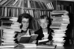 Two librarians from the Aurora Public Library in Aurora, Colorado, sit at a table, partially hidden by piles of books. Behind them are bookshelves full of more books. They each hold open books and book cards in their hands.