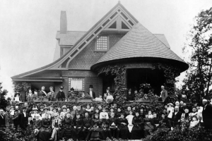 Portrait of well-dressed men, women, and children at a house in Denver, Colorado; costume includes bodices, flounces, straw, bowler, and stovepipe hats. The residence has shingle imbrication, a round terrace, and bargeboard.