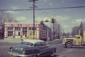 View of Gibbs Tire and Battery Company at 4589 Washington Street in the Globeville neighborhood of Denver, Colorado. An automobile and a truck make their way through the intersection of Washington Street and 46th Avenue. Signs on the building read: "Tires We Buy Sell Trade Tubes."