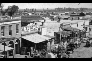 Horse-drawn wagons are parked near buildings on Blake Street in downtown Denver, Colorado. Signs on the buildings read: "Wholesale Groceries," "H. Burton Storage & Commission," and "Missouri House." The hotel has other signs: "Bar Room," "Boarding," and long horned cattle are tethered among the horses. Water of the South Platte River is in the background.
