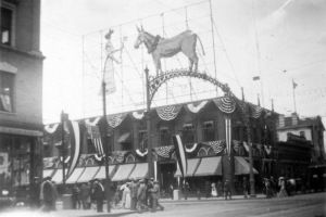Pedestrians stand near the Aspen Building (while it was home to the Gas and Electric Company) on the corner of 17th (Seventeenth) Street and Tremont Place in downtown Denver, Colorado. The building is decorated with bunting and has a lighted sign of a woman who holds a key out to a donkey on the roof. Lettering on the donkey reads: "Democracy." The electric sign is near a decorative metal arch that spans the street, both were erected for the Democratic National Convention.