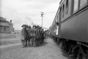Uniformed troops of infantry men board a Denver and Rio Grande train of Pullman sleeping cars, about to depart Fort Logan, a World War I assembly point for young soldiers, Colorado. Many wear uniforms consisting of jackets, jodhpurs, gaiters, boots and wide brim hats with Montana creases. A Black man, possibly a porter, stand in doorway of one of the cars. He wears a white coat and dark hat.